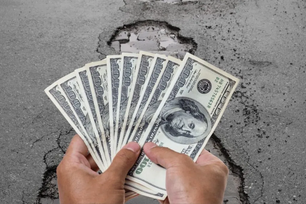 potholes on asphalt driveway and a hand holding dollar bills